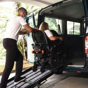 An aid pushes a person in a wheelchair up a ramp into a handicap-accessible van